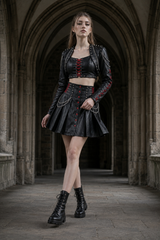Women’s gothic leather outfit in stone corridor with studded crop top, punk bolero jacket, red lacing, chain mini skirt, and boots - Rizwards Leather